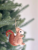 A charming wool felt squirrel ornament is suspended by a jute string against a softly blurred green Christmas tree. The brown squirrel features a lighter brown front, a white face, and a black eye. Its bushy tail is adorned with embroidered red berries and green sprigs, adding a cozy, rustic charm.