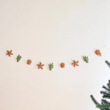 A wool felt starfish garland is displayed against a neutral wall, evoking a calm, coastal atmosphere. The garland features alternating tan felt starfish with dark brown stitching, green felt seaweed with dark green dots, and tan felt seashells with smiling faces. It is strung on a clear string and attached to the wall with a clear hook.