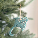 A handmade wool felt stingray ornament with a hemp string hangs on a festive green pine tree, creating a cheerful, artisanal atmosphere. The light teal stingray, featuring white polka dots, has a long curved tail and small black eyes, embodying an eco-friendly holiday spirit.