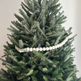 A white felt pompom ball garland gracefully drapes across a lush Christmas tree, creating a warm, celebratory atmosphere. The uniformly round, white felt pompoms are evenly spaced, providing a striking contrast against the vibrant green tree branches. This neat and polished garland enhances the natural texture of the foliage.