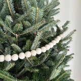 A Felt White Pompom Ball Garland made of wool felt is draped horizontally across the branches of a lush, green Christmas tree, evoking a cozy winter holiday atmosphere. The garland features evenly spaced, round white felt balls with a soft texture, nestled among the dark green evergreen needles, providing a gentle contrast.