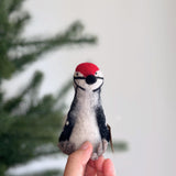 A vibrant handmade wool felt woodpecker finger puppet is gently held by a female hand, displayed against a blurred Christmas tree backdrop. This handcrafted felt kids toy features a vivid red crest, striking black and white body details, and a black beak. Its unique texture highlights its fair-trade craftsmanship.
