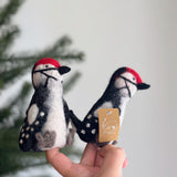A handmade felt woodpecker finger puppet, an eco-friendly creation, is held delicately in a female hand against a soft, festive backdrop with a glimpse of a Christmas pine tree. This needle felted puppet features a vibrant red crest atop its head, contrasting with its black and white patterned body. The handcrafted woodpecker showcases meticulous details, including a beak and distinctive markings, making it perfect for imaginative play.