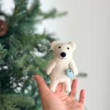 A handmade needle felted polar bear finger puppet, crafted from white wool felt and depicting a polar bear catching a small blue fish, is held in a female hand. The charming polar bear has small black eyes, a black nose, and a black mouth, displaying a cheerful expression. The blurred Christmas tree backdrop creates a festive and playful atmosphere for this unique finger puppet.