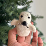 A handmade felt seal finger puppet, an endearing companion, is held in a hand against a blurred Christmas tree background. This off-white felt puppet features a black nose, whiskers, and eyes, along with flippers on each side. Its unique, handcrafted design makes it a charming toy for imaginative play.