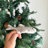 A playful felt shark finger puppet is held by a hand in front of a festive Christmas tree. This unique handmade shark puppet is light gray, featuring a dorsal fin, tail fin, and a distinct row of white teeth. It has black eyes and subtle stitches mimicking gills, making it an engaging felt toy for kids.