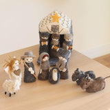 A handcrafted felt Christmas nativity scene, featuring a stable and eleven detailed wool felt finger puppets, is arranged on a wooden surface. The unique set includes Baby Jesus, Mary, Joseph, three kings, an angel, a shepherd, a fluffy white sheep, a dark gray and brown donkey, and a cow. The angel has golden yarn hair, and the kings, dressed in various grays and browns, each hold a golden box.