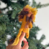 A fluffy wool felt lion finger puppet, handcrafted ethically in Nepal, is set against a blurred green Christmas tree, creating a festive scene. This unique felt toy features a bright yellow body, a fluffy mane with gradients of brown, tan, and gold, and black felt claws on its front paws.