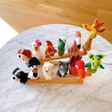 A playful collection of twelve handcrafted felt finger puppets is displayed on a light-colored bamboo stand, resting on a round marble table. Each unique wool felt finger puppet, including animals like a dog, duck with a flower, chicken, alligator, flamingo, giraffe, sheep, wolf, bird, and dragon, showcases vibrant colors and textures. This diverse handmade felt kids toy set offers engaging imaginative play.