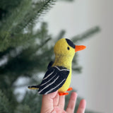 A handcrafted needle-felted goldfinch finger puppet is held gently by a female hand, with a blurred hint of a Christmas tree in the background. This charming wool felt finger puppet is predominantly vivid yellow with distinct black markings on its wings and head, complemented by a small orange beak and feet. Its detailed craftsmanship highlights the soft textures of the ethically made felt, perfect for imaginative play.