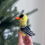 A handcrafted wool felt Goldfinch finger puppet is held by a female hand against a blurred Christmas tree. This vivid yellow and black needle-felted Goldfinch finger puppet features a distinct black patch on its head, black wings with white stripes, and a small orange beak.