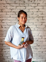 A wool felt goldfinch finger puppet is gently cradled in the hands of a Nepalese female artisan, wearing a light blue top, against a rustic white brick wall. This handcrafted felt goldfinch finger puppet features a vivid yellow body, black wings with white stripes, a black cap, and a small orange beak. Its delicate, needle-felted details capture the cheerful essence of the beloved songbird, highlighting its fair-trade craftsmanship.