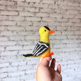 A handcrafted wool felt goldfinch finger puppet is held by a hand against a blurred white brick wall. This vibrant needle-felted goldfinch puppet features a bright yellow body, black head marking, orange beak and feet. Its black wings have white stripes, and the tail is tipped with black, highlighting the textures of this ethically made felt toy.