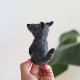 A needle felted gray coyote finger puppet, crafted from 100% wool, is gently held by a female hand against a neutral backdrop with a blurred green plant. This unique handmade felt finger puppet features a textured gray body, pointed ears, a tail, and striking blue eyes. Its detailed design highlights its artisanal craftsmanship.