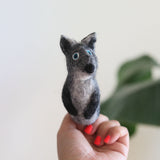 A detailed, handmade gray coyote finger puppet, crafted from 100% wool and needle felted, is held in a close-up by a female hand with red nail polish. The felt toy displays gray, white, and black colors, featuring pointed ears and small blue eyes for its 3D design. It rests against a softly lit, blurred backdrop of a white wall and a green plant, creating a calm, natural atmosphere.
