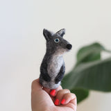 A handmade needle felted gray coyote finger puppet is playfully held by a female hand with red-painted nails, set against a bright backdrop with blurred green foliage. This handcrafted wool felt finger puppet showcases gray and white felt, small perky ears, a rounded black nose, and bright blue eyes, highlighting its unique design.