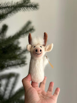 A handmade needle felted Highland Cow felt finger puppet is gently held by a female hand near a festive Christmas tree. Crafted from soft wool felt, this unique felt toy features a charming smiling face with a pink nose, black stitched details, fluffy white wool, and brown horns. A small tag is attached to its side, completing this delightful, handcrafted piece.