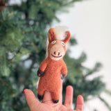 A light brown, needle-felted horse finger puppet made from 100% wool is held by a female hand against a blurred Christmas tree background. This unique horse features a white fluffy mane, brown ears, black hooves, and a stitched smiling face, ideal for engaging felt puppet play.