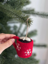 A red felt hot cocoa with marshmallow ornament hangs from a Christmas tree, with a blurred white wall in the background. The mug-shaped felt ornament features white snowflake patterns and is filled with brown felt mimicking cocoa, topped with small white felt marshmallows. A jute string allows it to hang gracefully.
