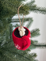 A handmade felt hot cocoa marshmallow ornament hangs from a green Christmas tree branch, set against a soft white background. The unique Christmas ornament features a red cup with white snowflake patterns, filled with brown felt cocoa and topped with fluffy white felted marshmallows. A jute string loop is attached for hanging, completing this charming Christmas tree decoration.