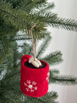 A hot cocoa with marshmallow felt ornament hangs on a Christmas tree, suspended by a jute string. This festive, handcrafted felt ornament is red and cylindrical, adorned with white snowflake patterns. An opening reveals dark brown felt inside, topped with white marshmallow-shaped pieces, creating a cozy and warm feel.