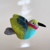 A vibrant wool felt hummingbird, part of a Hummingbird Baby Mobile, is suspended from a white felt frame by a thin white string against a light gray backdrop. This colorful felt hummingbird features a black beak, light green head, a pink and gray eye stripe, a blue belly, and green wings with a blue gradient, ending in a small gray tail. The soft felt material gives it a textured appearance.