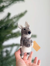 A handmade wool felt kangaroo finger puppet is gently held by a female hand, set against a soft, blurred green background suggesting a Christmas tree. This unique, fair-trade kangaroo features a larger gray and white figure carrying a smaller joey in its pouch, crafted with intricate needle-felted details. Made from 100% eco-friendly wool felt, this charming kids toy highlights traditional craftsmanship.