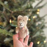 A handcrafted felt llama finger puppet is gently held by a female hand, appearing against a softly blurred Christmas tree with warm lights. The light brown llama features a sweet smile, soft fuzzy texture, white ear and foot details, and a charming pastel pompom collar.