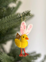 A whimsical handcrafted felt mini bunny chick ornament hangs from a Christmas tree branch. Its bright yellow chick body has an orange beak and detailed red felt legs. Long, soft white bunny ears with pink speckles top its head, complemented by a tan felt tail. It hangs from a yellow cotton string.