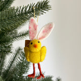 A whimsical mini needle felted bunny chick ornament hangs from a pine tree branch against a soft white backdrop, evoking a playful Easter mood. The charming yellow bunny chick features a round body, an orange beak, black eyes, and long, soft pink ears. Thin red legs and a jute string complete this unique handmade felt ornament.