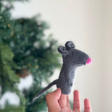 This handcrafted grey felt rat finger puppet is playfully held by a fair-skinned hand, set against a blurred background of a festive green Christmas pine tree and a bright white wall. Crafted from soft wool felt, this unique rat finger puppet features small rounded ears, a pink nose, and a thin tail, inviting imaginative storytelling and play.