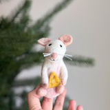 A female hand gently holds a unique needle felted white and pink mouse holding cheese finger puppet, presented against a soft, blurred backdrop of a festive Christmas tree. The charming mouse features tiny black eyes, a small pink nose, and delicate white whiskers, expertly clutching a small, triangular piece of yellow cheese. This handcrafted wool felt mouse finger puppet, showcasing detailed needle felting, embodies the festive spirit and fair-trade craftsmanship.