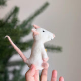 A handcrafted wool felt mouse finger puppet, holding a small yellow piece of cheese, is delicately held by a female hand. The white mouse features pink accents on its ears, paws, and tail. In the blurred background, a Christmas tree suggests a festive atmosphere. This unique felt toy showcases a soft, textured appearance and detailed craftsmanship, making it a charming and compact puppet.