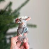 A unique handmade wool felt mouse finger puppet holding a light blue cheese is presented in a female hand against a blurred Christmas tree background. This grey needle-felted mouse features a white hat, tan ears, a pink nose, and white whiskers. Crafted with fair-trade principles, it’s an eco-friendly felt toy for kids.