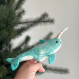 A needle felted narwhal finger puppet is held by a female hand against a soft, festive backdrop with a blurred Christmas tree. This handcrafted felt kids toy, made from soft light blue and white wool felt, features a gentle smile, fins, and a distinctive spiraled tusk.