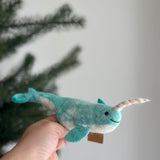 A handmade wool felt narwhal finger puppet is presented by a female hand against a blurred Christmas pine tree backdrop. This unique blue narwhal features white markings, tiny black eyes, a small stitched smile, its signature spiraled tusk, and matching blue fins and tail.