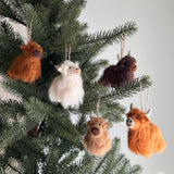 Needle felted Highland Cow ornaments are displayed on a lush green Christmas tree, set against a soft, neutral background. Each charming ornament, suspended by a jute string, features soft fluff, cute horns, and delicate needle-felted details, including a stitched smile. These handcrafted felt ornaments come in various colors such as brown, white, and orange, creating a warm, playful, and festive display.