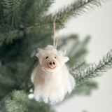A needle felted Highland Cow Christmas ornament hangs playfully on a green Christmas tree. This unique Christmas ornament is mostly white with soft fluff, cute horns, a pink snout, a stitched smile, and small black eyes. A brown jute string is attached for easy hanging, adding a soft and charming touch.