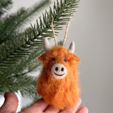 A needle felted highland cow ornament is shown in two views: gently held in a female hand and hanging from a green Christmas tree branch, evoking warmth and festive cheer. This handcrafted felt ornament features a fluffy orange body with delicate needle-felted details, black eyes, and cream-colored horns. A jute string is attached for hanging.