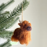 A needle felted highland cow ornament hangs from a lush green Christmas tree branch, creating a cozy and festive atmosphere. This charming handcrafted felt ornament features a soft brown rounded body, a dark brown nose, and small white horns, suspended by a jute string.