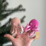 Two soft, handcrafted wool felt octopus finger puppets are playfully displayed on a female hand against a festive Christmas tree. One is light pink, the other magenta, each featuring eight tiny curled tentacles, poppy smiley eyes, and a charming grin. The magenta felt octopus finger puppet also has a small tag, completing the cheerful scene.