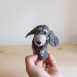 A handcrafted wool felt old gray dog finger puppet, made by female artisans in Nepal, is presented by a female hand with red nail polish. The charming gray felt puppet, featuring long floppy ears and a light pink nose, rests against a soft white backdrop and a light wood surface, creating a calm atmosphere.