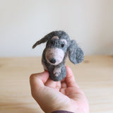 A handmade needle felted old gray dog finger puppet is held by a female hand against a light beige background. Crafted from 100% wool in Nepal, this unique 3D puppet is predominantly gray with distinctive floppy ears, a pinkish-gray nose, and small dark eyes, showcasing its artisanal texture.