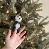 A handcrafted felt panda finger puppet is held upright against a warm, lit Christmas tree. This black and white panda features a round face, plump body, black ears, and eye markings that distinctively contrast with its white face and belly. The charming felt toy is ready for imaginative play.