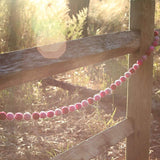 A Pink Felt Ball Garland, crafted from soft wool felt, hangs gracefully on a weathered wooden fence. It features round balls in various shades of pink, bathed in warm, golden sunlight filtering through tall grasses and trees. The outdoor setting creates a charming and inviting atmosphere.