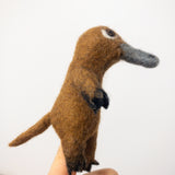 A handmade brown platypus felt finger puppet, crafted from 100% wool, is delicately held on a female finger against a stark white backdrop. This unique felted finger puppet features a distinctive grey bill, small round eyes, and detailed flippers, showcasing its meticulous needle-felted craftsmanship and playful appearance.