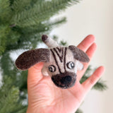 A charming pug felt finger puppet is gently cradled in a female hand, set against a blurred Christmas pine tree. This light beige felt finger puppet features darker brown vertical stripes on its head, floppy dark brown ears, and a black nose. Detailed with expressive black and white felt eyes, this handcrafted felt toy for kids showcases meticulous craftsmanship.