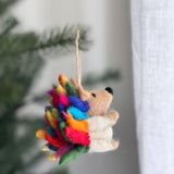 A vibrant rainbow hedgehog ornament is suspended on a Christmas tree, set against a soft, blurred background. This wool felt ornament features bright rainbow-colored spikes, transitioning from green, red, orange, yellow, blue, and magenta, with a tan face and body, tiny black nose, and ears. It hangs by a rustic jute string, adding a joyful, festive touch.