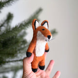 A handmade red fox wool felt finger puppet is gently presented by a female hand against a soft white backdrop with a hint of a Christmas tree. This charming, meticulously crafted felt fox puppet features a white belly, inner ears, and muzzle. Black accents on its feet, nose, and ear tips, along with delicate whiskers, complete its endearing design.
