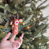 A festive needle felted Red Reindeer Finger Puppet rests on a female hand, with a blurred Christmas tree and yellow lights in the background creating a warm holiday setting. This charming felt reindeer puppet is primarily red and cream, featuring brown antlers and a distinct red nose.